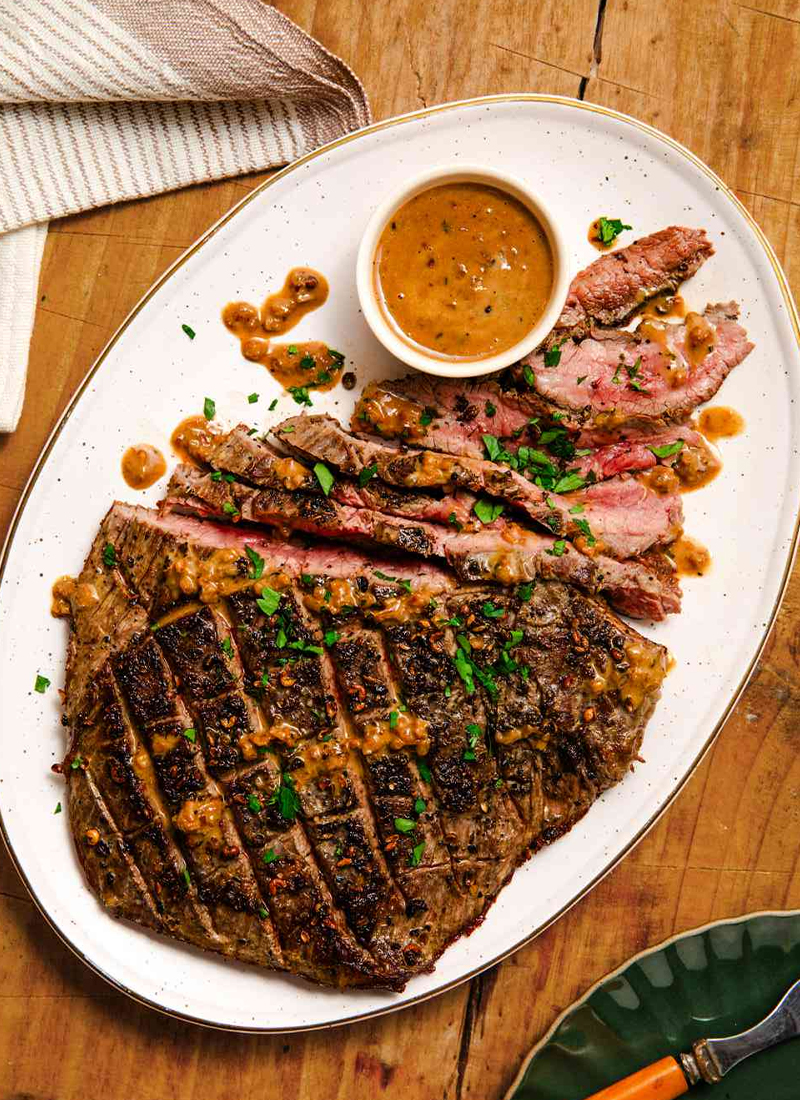 Sliced grilled steak served with peppercorn sauce on a rustic white plate, garnished with fresh herbs served at Mahony’s Steak House Bishopbriggs.